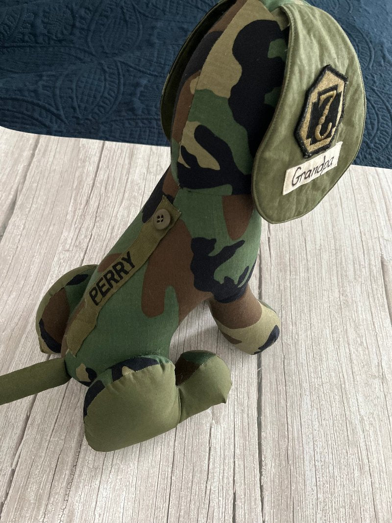 A keepsake military dog made from an upcycled army coat is sitting on a bed. The dog has an army medic badge sewn on one ear.