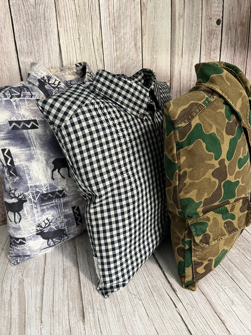 Three keepsake pillows standing beside each other. Pillows are made from customers clothes. One pillow has moose moif in the fabric design, another is black and white gingham check, and the last is brown, green camouflage design. Each pillow measures 16x16 inches.