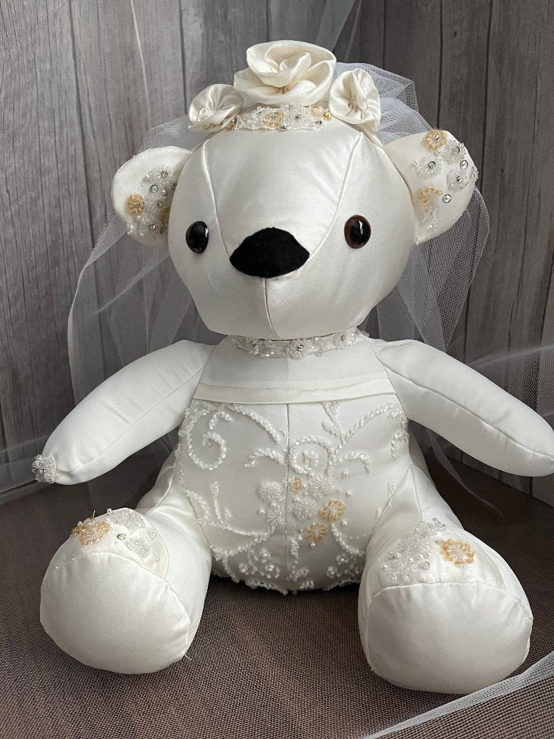 White keepsake wedding bear made from customers wedding gown. The bear sits at 12 inches tall and weighs 1.5 pounds. It has lace and pearls stitched to the bodice back and front and on the sole of the feet. It has a veil and three bows on the head, black safety eyes and black felt nose.