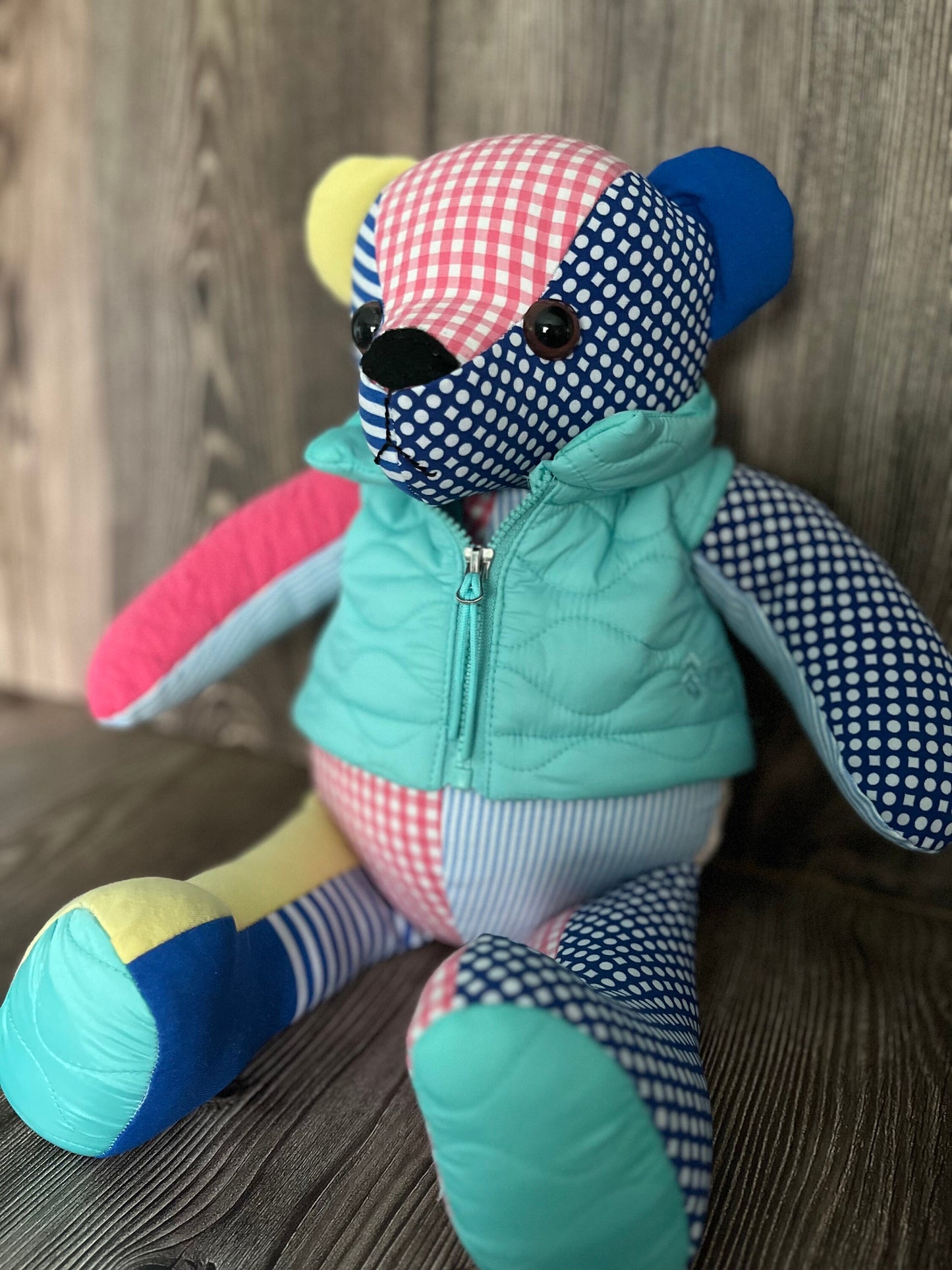 18 inch memory bear made from 6 upcycled clothes. The bear is sitting and wearing a light blue zipped up vest. Colors are light blue, pink, blue, and yellow.