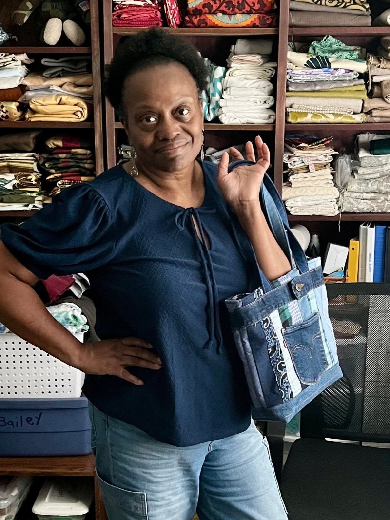 Black woman wearing jeans and short sleeved blouse holding a denim tote made with strips of quilted fabric made from upcycled men's shirt and denim pants.